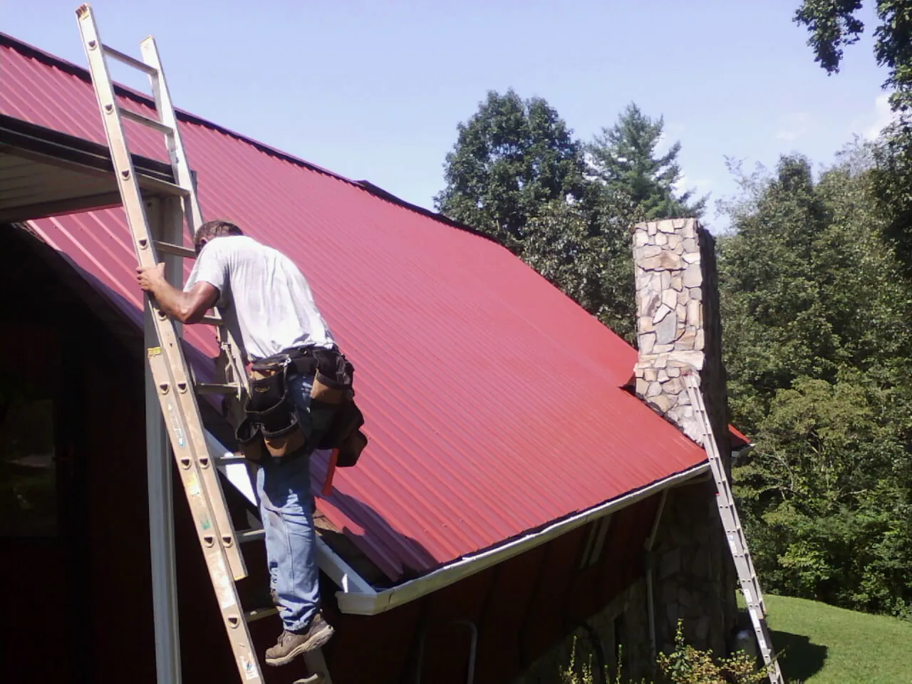 Roofer on a ladder beside a red metal roof and stone chimney — Soffit & Fascia Repair by Roofing Brothers in Burnsville